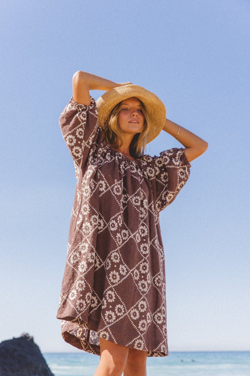Dahlia Mini Dress Maple — side angle model at beach, arm raised to straw hat, blue sky background, hand block print linen mini in maple brown geometric pattern, slow fashion artisan dress | Daughters of India