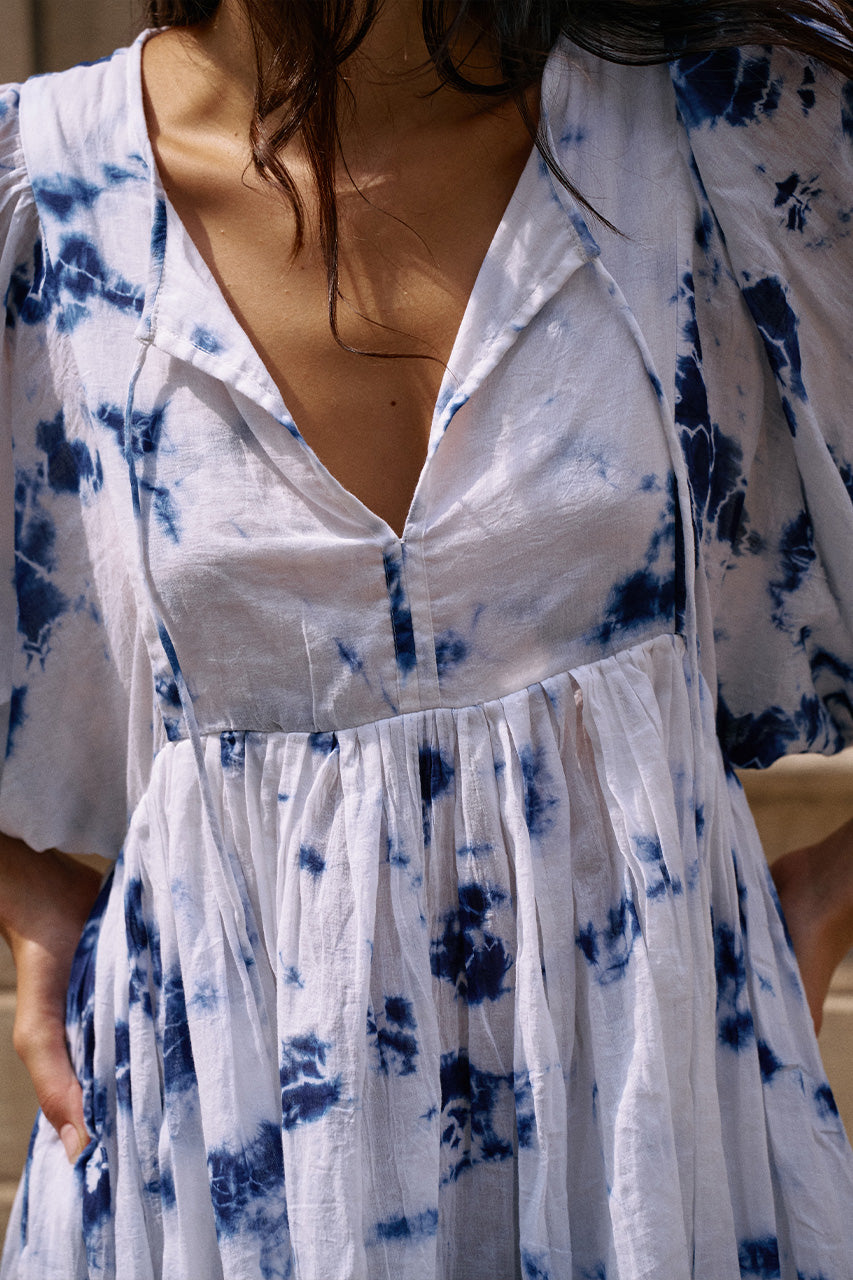 Kyra Mini Dress Deep Sea — full length side view walking on sidewalk, deep sea shibori tie-dye cotton mini dress with flowing skirt movement, stone building facade | Daughters of India